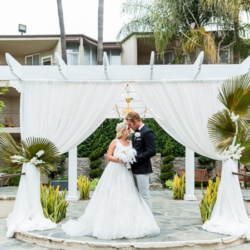 A couple in wedding attire share a kiss under a white draped arch on a circular stone platform with palm plants and greenery around.