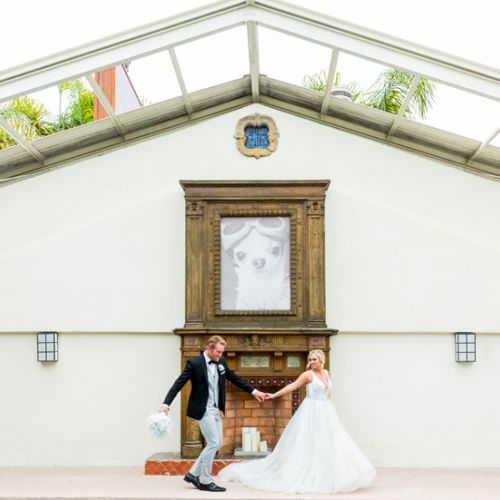 A bride and groom in wedding attire pose outside a white building with an archway and ornate doorway.