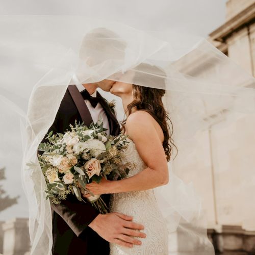 A bride and groom kiss outdoors, the bride holding a bouquet of flowers under a flowing wedding veil.