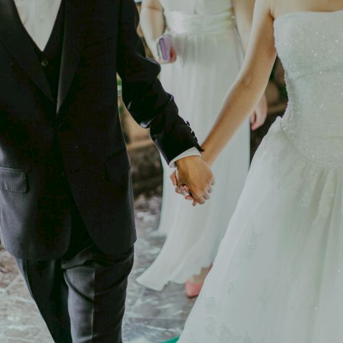 A bride and groom hold hands in wedding attire, captured waist-down during their ceremony.