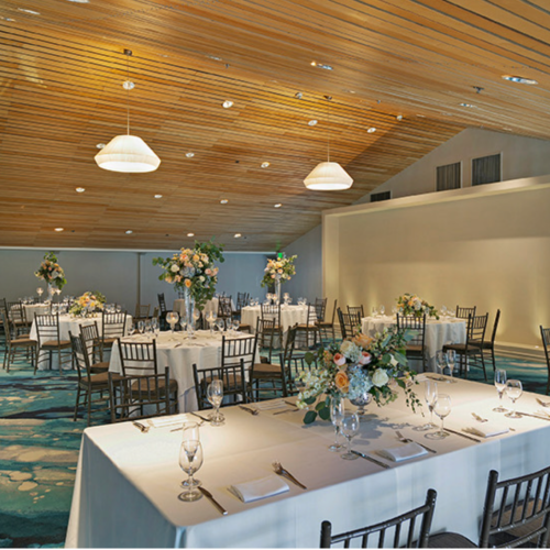 A decorated banquet hall set for a wedding reception, with long tables, candles, floral centerpieces, and hanging pendant lights.