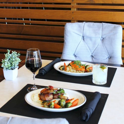 A dining table setup with two plated entrees, red wine and water glasses, folded napkins, and black place mats.