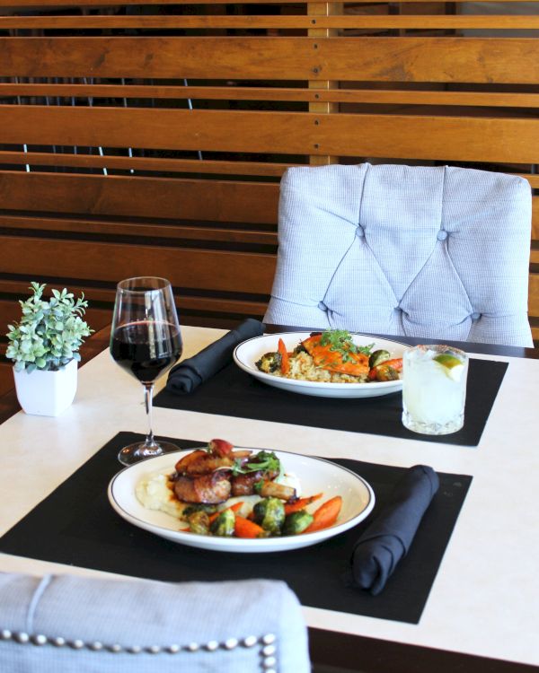 A dining table setup with two plated entrees, red wine and water glasses, folded napkins, and black place mats.