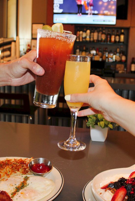 Two people clink tall frosty cocktails&mdash;one red, one yellow&mdash;at a bar table, with pizza and dessert plates in view.