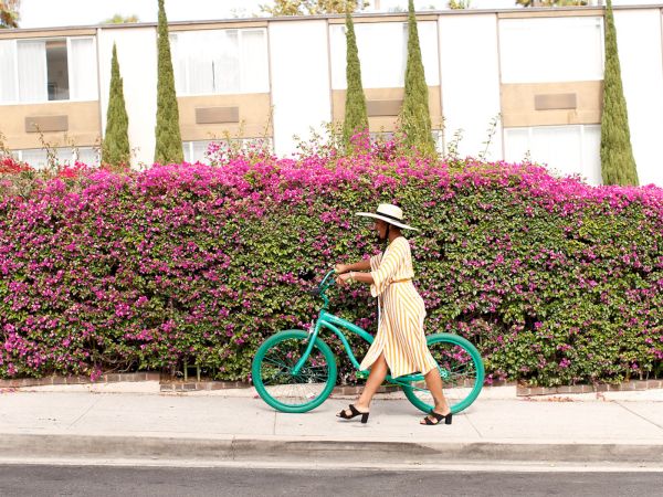 A woman in a striped dress rides a turquoise bicycle past a building covered in colorful pink flowers.