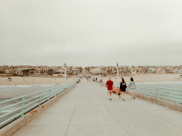 People stroll on a seaside boardwalk beside waves, with a coastal town and palm trees stretching along the beach.