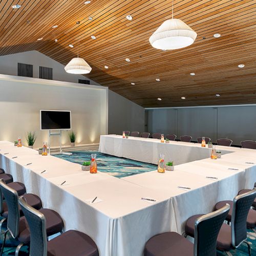 A square U-shaped conference table setup around a water feature in a bright, wood-ceiling room with a wall TV, plants, and many chairs.