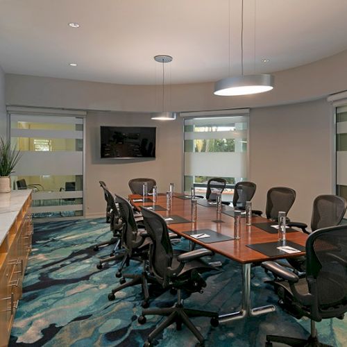 Modern conference room with a long wooden table, mesh swivel chairs, blue carpet, and large window blinds.