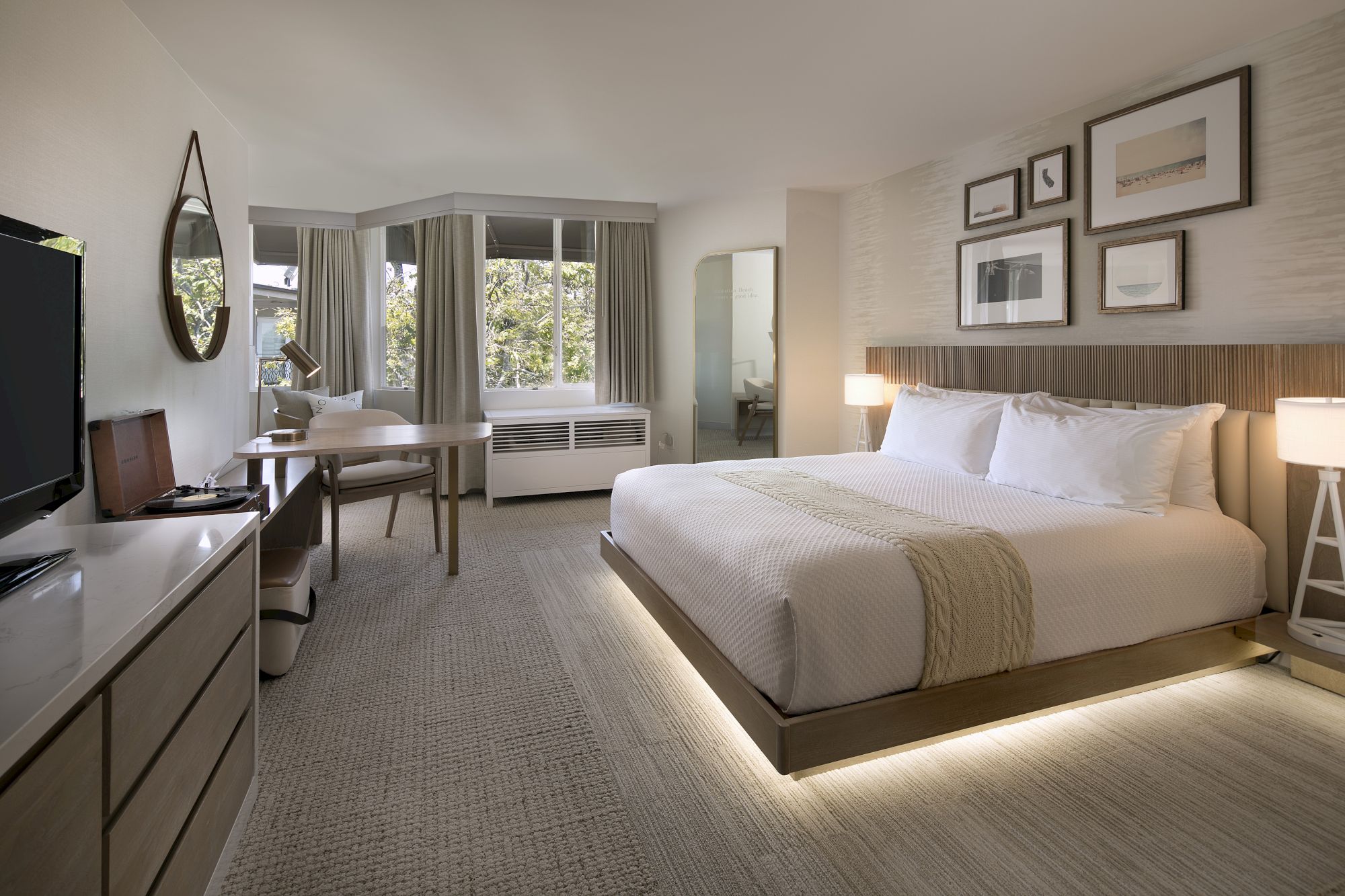 A modern hotel room with a neatly made bed, TV and dresser, desk, and sunlit windows with curtains and framed art.