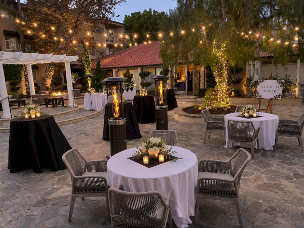 An outdoor &ldquo;Rehearsal Dinner&rdquo; patio setup with string lights, candlelit tables, wicker chairs, and two tall flame heaters.