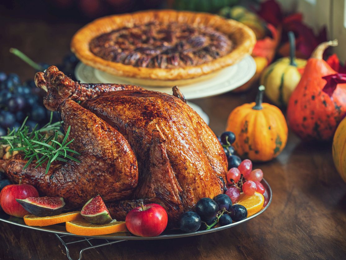 Roasted turkey on a platter with fruits and herbs, plus a baked pie in the background.