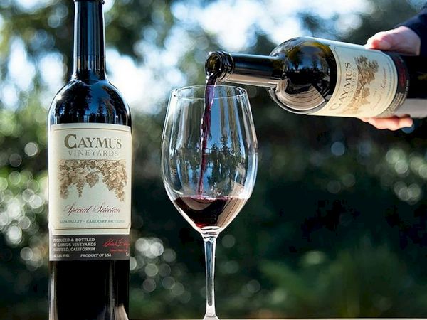 A hand pours red wine from a bottle (Caymus Vineyards) into a glass with a smoky bokeh background.