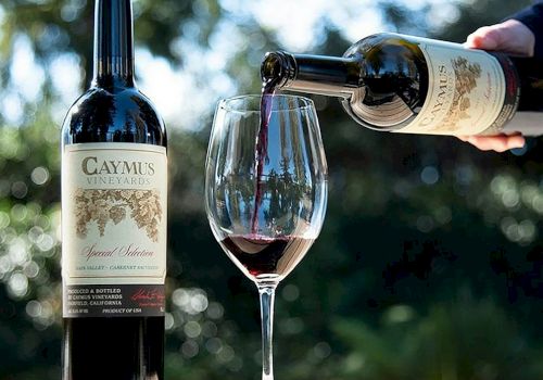 A hand pours red wine from a bottle into a glass beside a CAYMUS VINEYARDS bottle, with a smoky bokeh background.