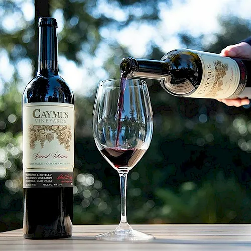 A hand pours red wine from a bottle into a glass beside a CAYMUS VINEYARDS bottle, with a smoky bokeh background.