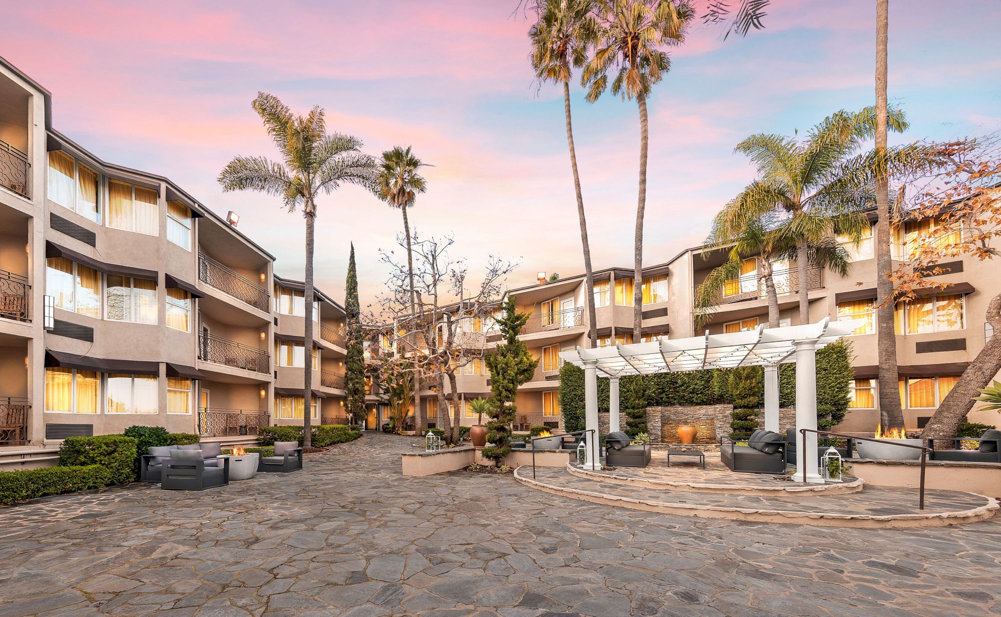 A hotel courtyard at sunset with palm trees, beige multi-story rooms, and a white pergola over outdoor seating and fire pits.