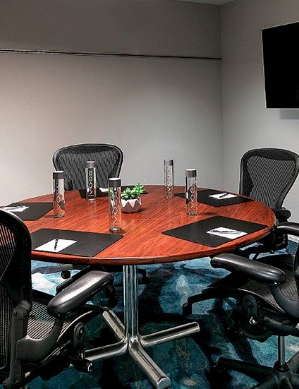 A modern meeting room with a round wooden table, mesh office chairs, water carafe, and a wall-mounted TV.