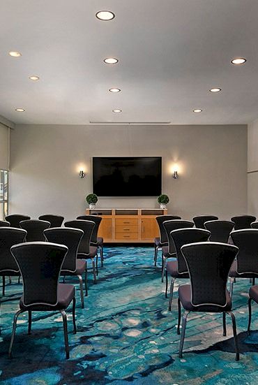 A conference room with rows of upholstered chairs, a blue carpet, and a large wall TV at the front.