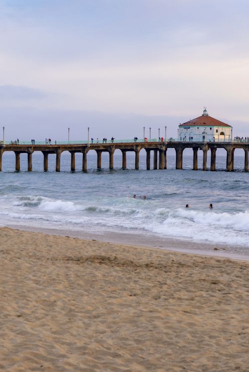 A seaside pier with a lighthouse stands over the ocean as waves roll in under a pastel sunset sky.