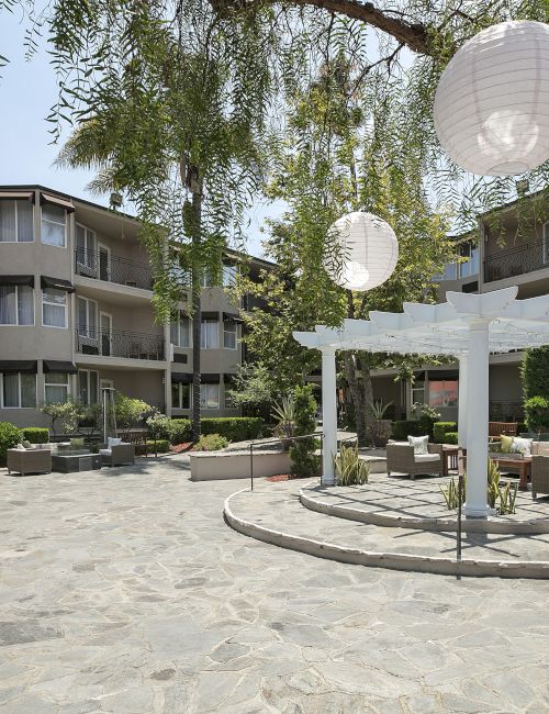 A sunny hotel courtyard with seating, a pergola, circular lantern-like hanging lamps, and lush trees above apartment balconies.