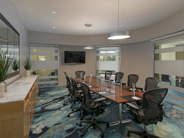 Modern conference room with a long table, swivel chairs, wall TV, and blinds-covered windows, lit by ceiling pendant lights.