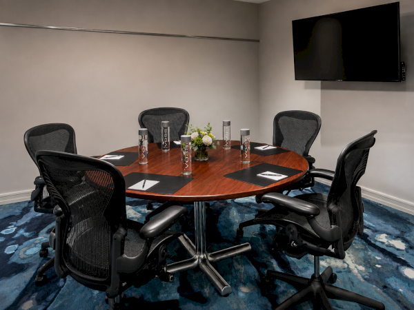 Meeting room with a round wooden table, ergonomic chairs, bottled drinks, flowers, and a wall TV above blue carpet.