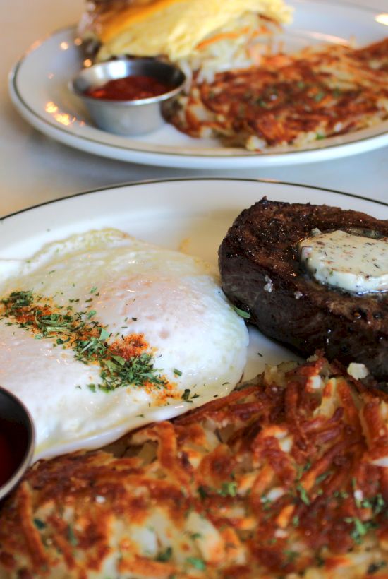 Plates of hearty breakfast: a steak with fried egg, pizza slices, veggie salad, and small bowls of sauce.