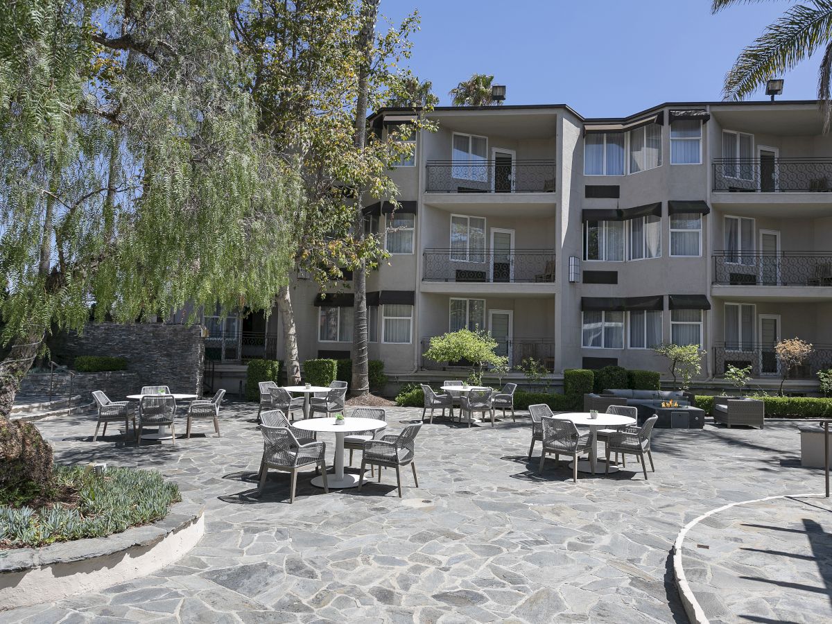 Sunlit apartment courtyard with patio chairs and tables, a fire pit, tall trees, and balconies under a clear blue sky.