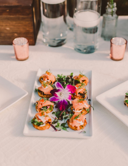 A catered table with floral garnished shrimp appetizers on white plates and glasses of water behind them.