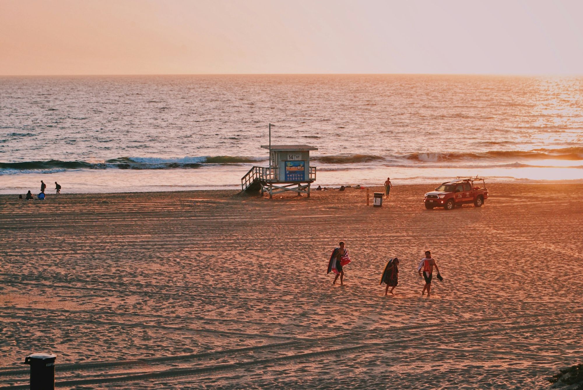 Sunset at the beach with a lifeguard tower, waves rolling in, a few walkers, and a parked pickup near the shore.