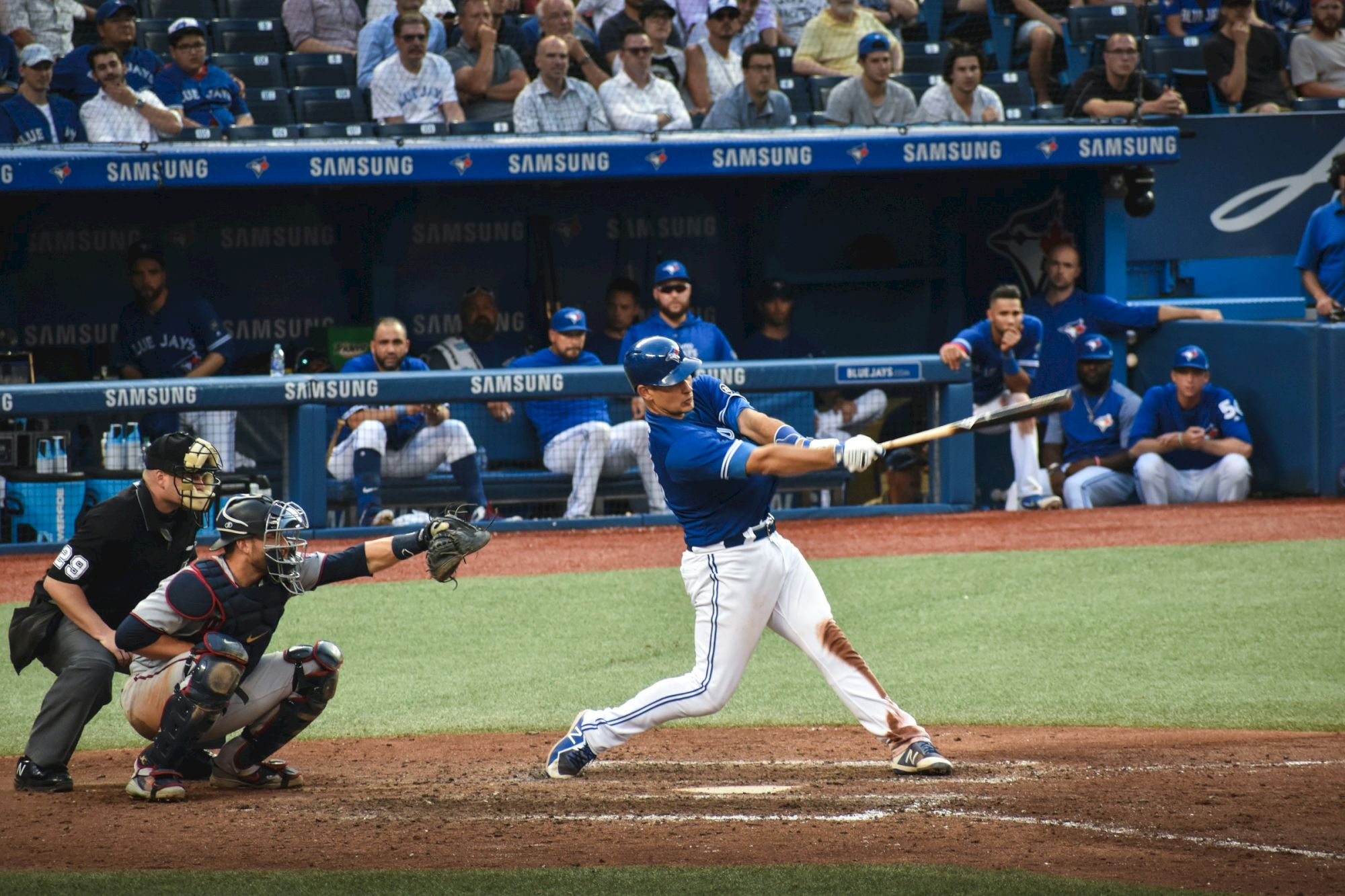 A Toronto Blue Jays batter swings at a pitch as the catcher and umpire crouch behind home plate in a packed stadium.