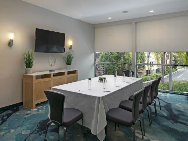 Modern conference room with a wall TV, wall sconces, motorized window shades, and a long table set for meetings, overlooking greenery.
