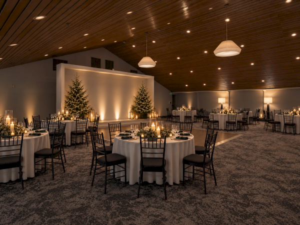 A warmly lit banquet hall set for a holiday dinner, with round tables, candles, and glowing Christmas trees.