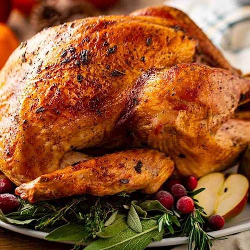 A roasted, golden-brown whole chicken with herbs on a platter, surrounded by fruit and vegetables for a festive meal.